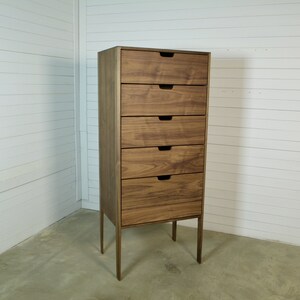 May include: A tall wooden dresser with five drawers and a light brown finish. The dresser has a simple design with metal legs.