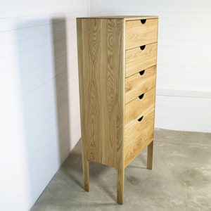 May include: A tall wooden chest of drawers with six drawers and black handles. The dresser has a light wood finish and stands on four legs.