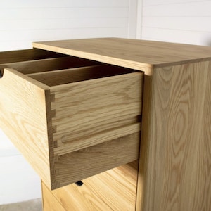 May include: A wooden chest of drawers with five drawers, the top drawer is open and reveals a dovetail joint. The dresser is made of light wood with a natural finish.