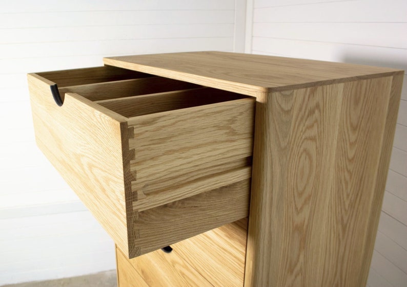 May include: A wooden chest of drawers with five drawers, the top drawer is open and reveals a dovetail joint. The dresser is made of light wood with a natural finish.