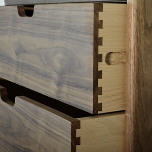 May include: Close-up of two drawers on a wooden dresser. The top drawer is open, revealing a dovetail joint and a light wood interior. The drawers are made of dark wood with a natural grain pattern.