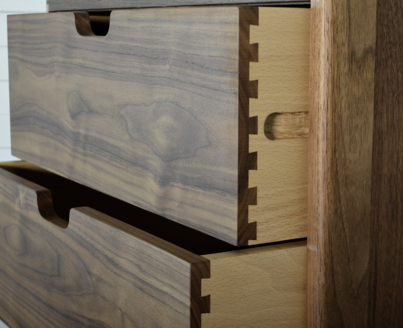 May include: Close-up of two drawers on a wooden dresser. The top drawer is open, revealing a dovetail joint and a light wood interior. The drawers are made of dark wood with a natural grain pattern.