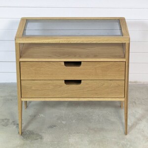 Puede incluir: Una mesita de noche de madera clara con dos cajones y una tapa de vidrio. La mesita de noche tiene un dise&ntilde;o simple y moderno y es perfecta para un dormitorio.