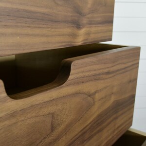 May include: Close-up of a wooden dresser drawer with a recessed handle. The drawer is made of a rich brown wood with a natural grain pattern.