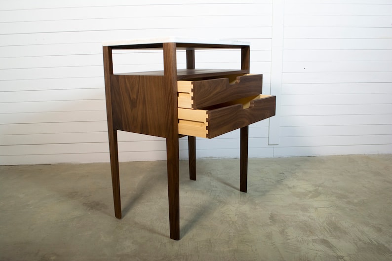 May include: A two-drawer bedside table with a walnut wood frame and a white marble top. The drawers are open, revealing a light wood interior.