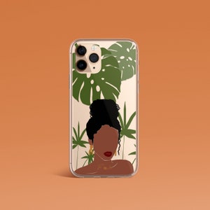 May include: A clear phone case with a design of a woman with dark brown skin and a bun, surrounded by green tropical leaves. The case has a gold camera ring and a gold chain with the letters "T.M.K." on it.