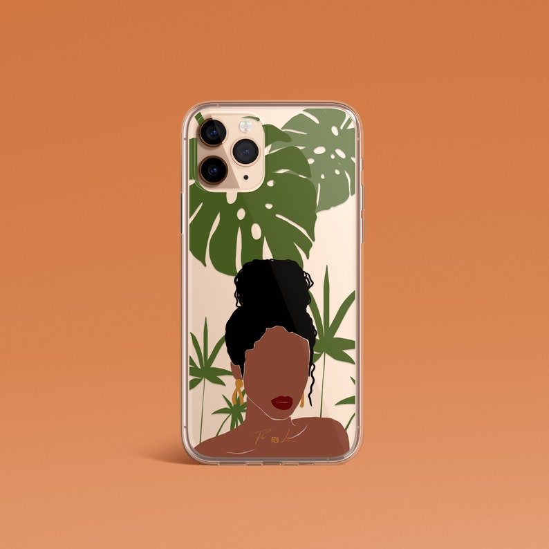 May include: A clear phone case with a design of a woman with dark brown skin and a bun, surrounded by green tropical leaves. The case has a gold camera ring and a gold chain with the letters "T.M.K." on it.