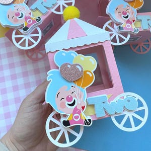 Puede incluir: Tren de circo de papel rosa con un payaso de dibujos animados sosteniendo globos. El tren tiene ruedas blancas y la palabra "Two" en el lateral. El payaso tiene el pelo azul y una nariz roja. El tren está decorado con un pompón amarillo.