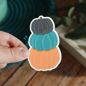 May include: A sticker featuring three stacked pumpkins in shades of orange, teal, and gray. The pumpkins have a white border and are held by a hand. The background includes a teal mat and a wooden surface.