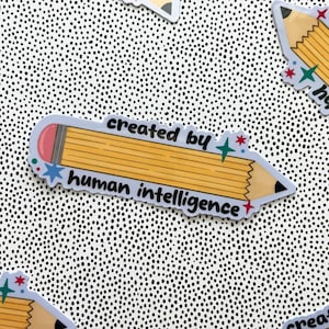 May include: A yellow pencil-shaped sticker with a pink eraser and black lead tip. The text "created by" is printed above the pencil and "human intelligence" below it, accented by small blue, red, and green star-shaped icons on the edges.