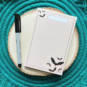 May include: A blank notecard with a light pink background and black bats printed on it. The notecard is sitting on a wooden surface with a teal fringe around it.