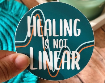 May include: A teal round sticker with a white outline and a white graphic of a wavy line. The text "Healing is not linear" is written in white on the sticker.