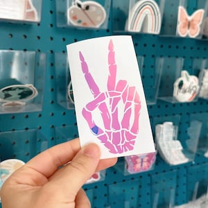 May include: A white sticker featuring a skeleton hand in a peace sign gesture, with a gradient of pink and purple hues. The sticker is held in front of a teal pegboard with various other stickers.