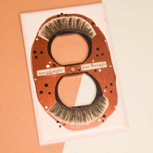 Pode incluir: Uma tampa de interruptor de luz branca com duas tomadas pintadas com um design de cobre e preto. A tomada superior tem o texto "Just enough" e a tomada inferior tem o texto "out-of-sight".