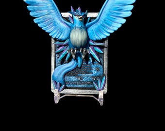 May include: A blue and purple Pokemon card featuring a blue bird with outstretched wings. The card is framed in a silver frame with a clear plastic stand.