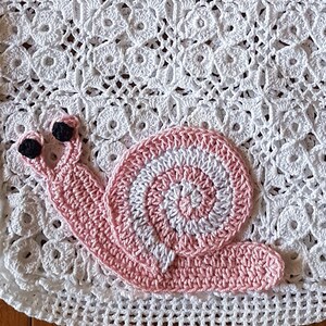 Peut inclure: Sac fourre-tout en crochet blanc avec un motif d'escargot. Le sac présente un motif ajouré détaillé et des anses robustes. L'escargot est rose et blanc avec des yeux noirs, ajoutant une touche fantaisiste.