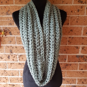 以下が含まれることがあります： 黒いマネキンに飾られた、セージグリーンのインフィニティスカーフ。スカーフは手編みで、テクスチャードステッチでリブ編みの模様を作り出しています。柔らかく落ち着いた色合いと居心地の良いデザインは、肌寒い季節にぴったりの暖かいアクセサリーです。
