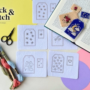 May include: Printable Stick & Stitch tea bag bookmark designs featuring white fabric sheets with printed patterns of stars, flowers, hearts, and moons, alongside embroidery floss, black scissors, and finished felt bookmarks on an open book.