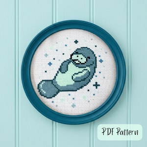 May include: A cross-stitch pattern of a manatee in shades of blue and green, set in a round teal frame. The manatee is surrounded by small blue crosses and stars. The text "PDF Pattern" is in a white rectangle.