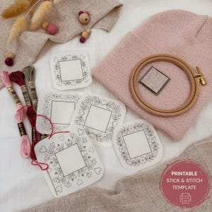 Könnte beinhalten: Eine rosa Mütze, ein Stickrahmen und mehrere weiße, bedruckbare Stick- und Stichvorlagen mit Blumen- und Herzmotiven sind auf einer weißen Oberfläche angeordnet. Stickgarn in Rosa- und Grüntönen ist ebenfalls vorhanden. Eine Vorlage mit dem Text "PRINTABLE STICK & STITCH TEMPLATE" ist sichtbar.