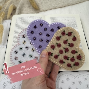 May include: Heart-shaped felt bookmarks in lavender, beige, and white, adorned with embroidered flowers, butterflies, and roses. The bookmarks are held over an open book. The image includes the text "NOTE: DMC COLORS & STITCH GUIDE INCLUDED".