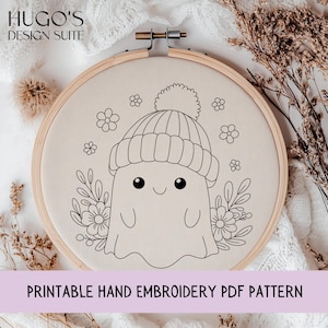 Puede incluir: Una tela beige estirada dentro de un bastidor de bordado de madera presenta un dibujo lineal negro de un fantasma con un gorro de punto. El fantasma está rodeado de diseños florales y de hojas. El texto "PRINTABLE HAND EMBROIDERY PDF PATTERN" está en la parte inferior.