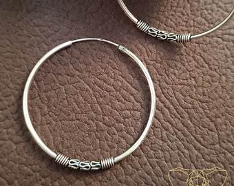 May include: Two silver hoop earrings with decorative detailing. The earrings have a simple, circular design with a series of small, ornate accents near the bottom. The earrings are displayed on a brown leather surface with a piece of driftwood in the background.