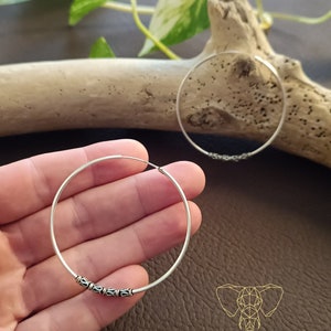 May include: A pair of silver hoop earrings with a decorative design. The earrings are shown on a hand against a brown background.