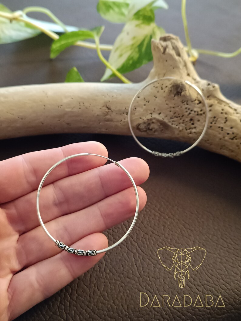 May include: A pair of silver hoop earrings with a decorative design. The earrings are shown on a hand against a brown background.