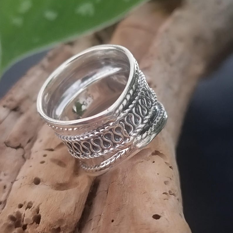 May include: A silver ring with an intricate woven design. The ring is wide and has a rounded band.