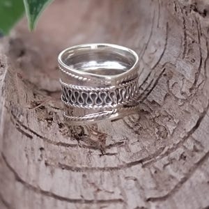 May include: A silver ring with a wide band and intricate woven detailing. The ring is made of sterling silver and has a polished finish.