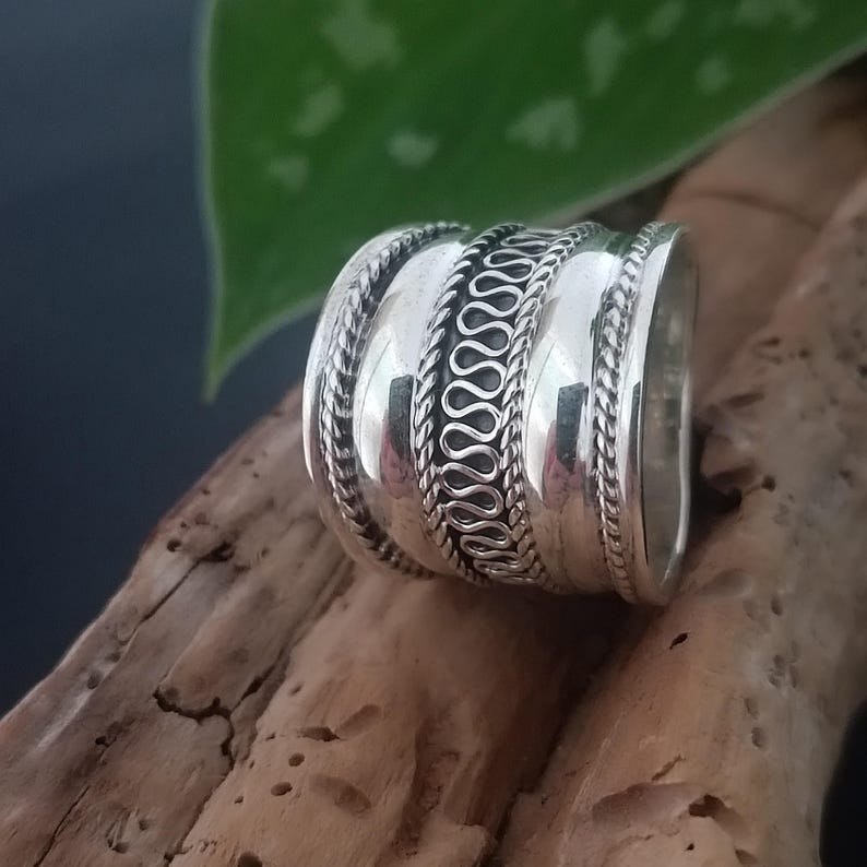 May include: A silver ring with a wide band and intricate designs. The band is decorated with three rows of raised, textured patterns.