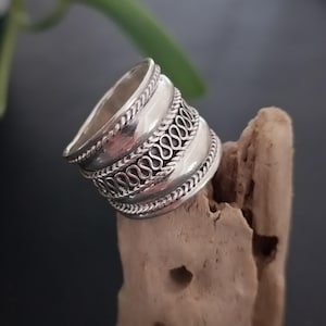 May include: A silver ring with a wide band and intricate designs. The band is decorated with three rows of repeating patterns, creating a textured and ornate look.