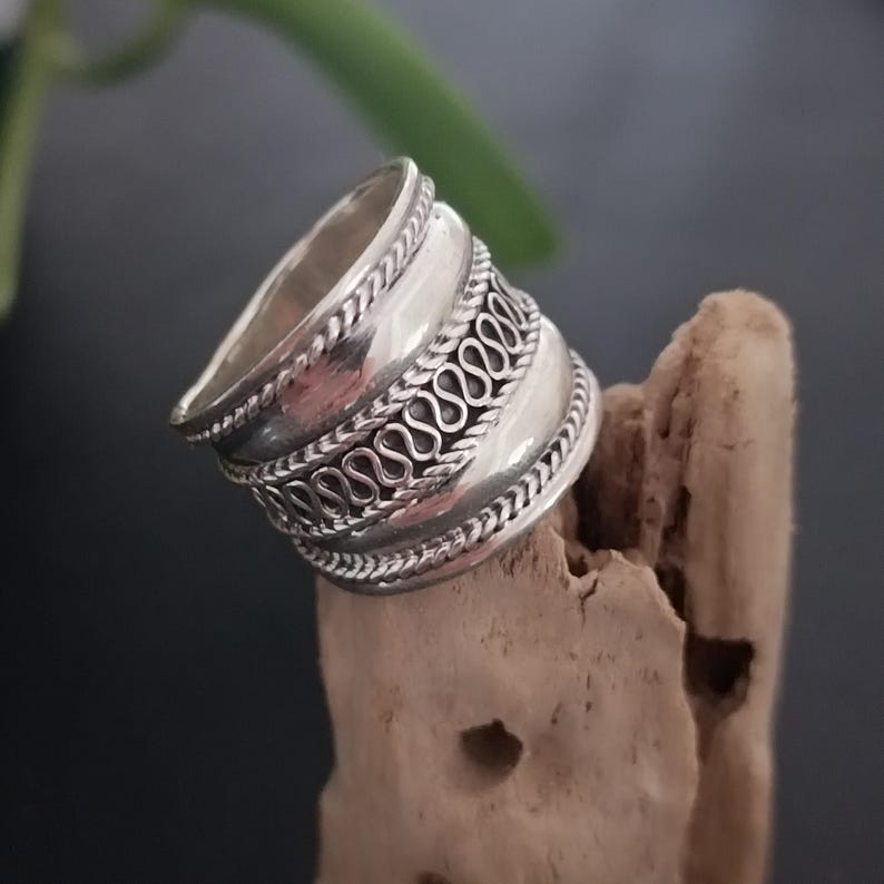May include: A silver ring with a wide band and intricate designs. The band is decorated with three rows of repeating patterns, creating a textured and ornate look.
