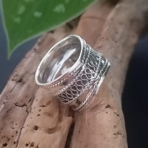 May include: A silver ring with a wide band and intricate woven design. The band is textured and has a slightly rounded shape.