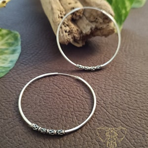 May include: Two silver hoop earrings with a decorative design of small circles around the hoop. The earrings are on a brown textured surface.