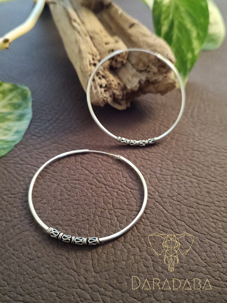 May include: Two silver hoop earrings with a decorative design of small circles around the hoop. The earrings are on a brown textured surface.