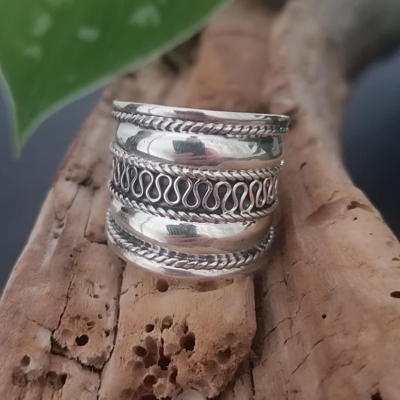 May include: A silver ring with a wide band and intricate designs. The band is made up of multiple layers, each with a different pattern. The ring is a statement piece and would be a great addition to any jewelry collection.