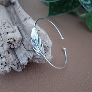 Könnte beinhalten: Ein silberfarbenes Armreif-Armband mit einem detaillierten Federdesign. Das offene Armband liegt auf einem Stück Treibholz vor einem braunen Hintergrund. Die Feder ist der Blickfang mit aufwendigen Details.