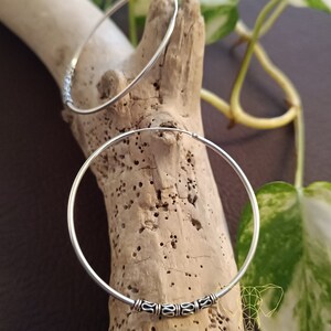 May include: Two silver hoop earrings with a decorative design on the front. The earrings are on a wooden surface.