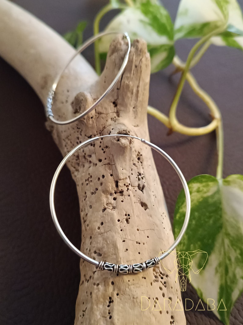 May include: Two silver hoop earrings with a decorative design on the front. The earrings are on a wooden surface.