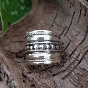 May include: A silver ring with a wide band and three rows of decorative detail. The top row features a rope-like design, the middle row has small beads, and the bottom row has a rope-like design.
