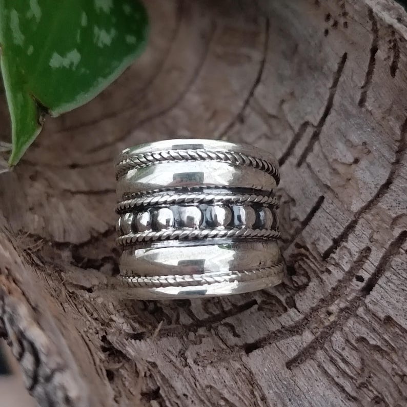 May include: A silver ring with a wide band and three rows of decorative detail. The top row features a rope-like design, the middle row has small beads, and the bottom row has a rope-like design.