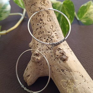 May include: Two silver hoop earrings with a decorative design on the bottom. The earrings are on a wooden surface.