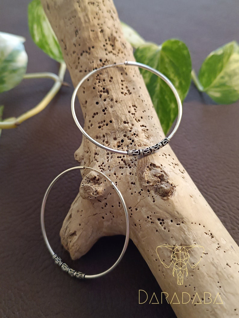 May include: Two silver hoop earrings with a decorative design on the bottom. The earrings are on a wooden surface.