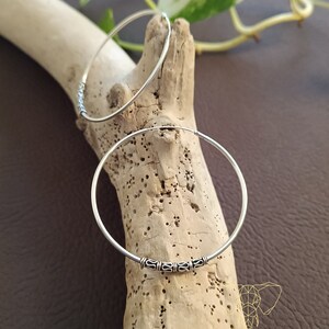 May include: Two silver hoop earrings with a decorative design on the front. The earrings are on a wooden surface.