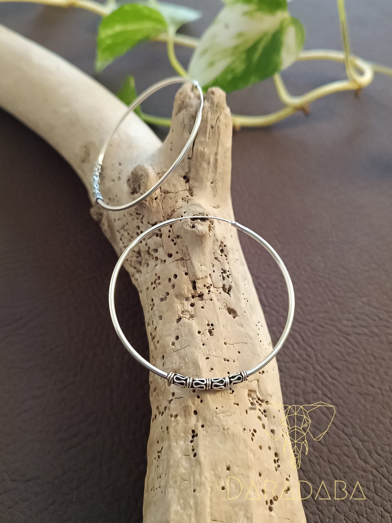 May include: Two silver hoop earrings with a decorative design on the front. The earrings are on a wooden surface.