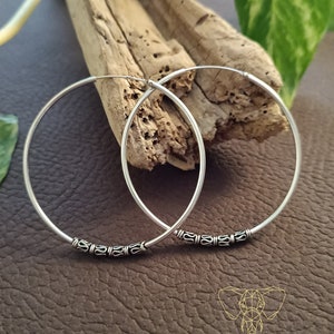 May include: A pair of silver hoop earrings with a decorative design around the edge. The earrings are on a wooden surface.