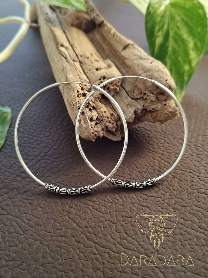 May include: A pair of silver hoop earrings with a decorative design around the edge. The earrings are on a wooden surface.