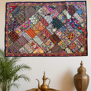 Op de afbeelding: Een levendig patchwork textiel wandkleed met een zwarte rand. Het ontwerp toont een verscheidenheid aan kleurrijke, geometrische patronen. De kamer is ingericht met een kleine houten tafel, kussens en een messing theeservies, wat een bohemien esthetiek creëert.